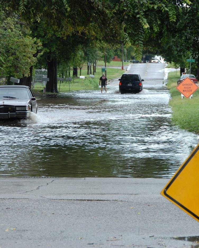 Cars driving slowly through a flooded street past a high water sign