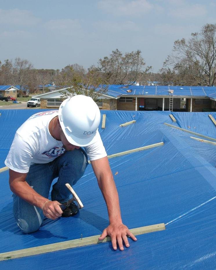 Roofer nailing down a blue tarp over a damaged roof
